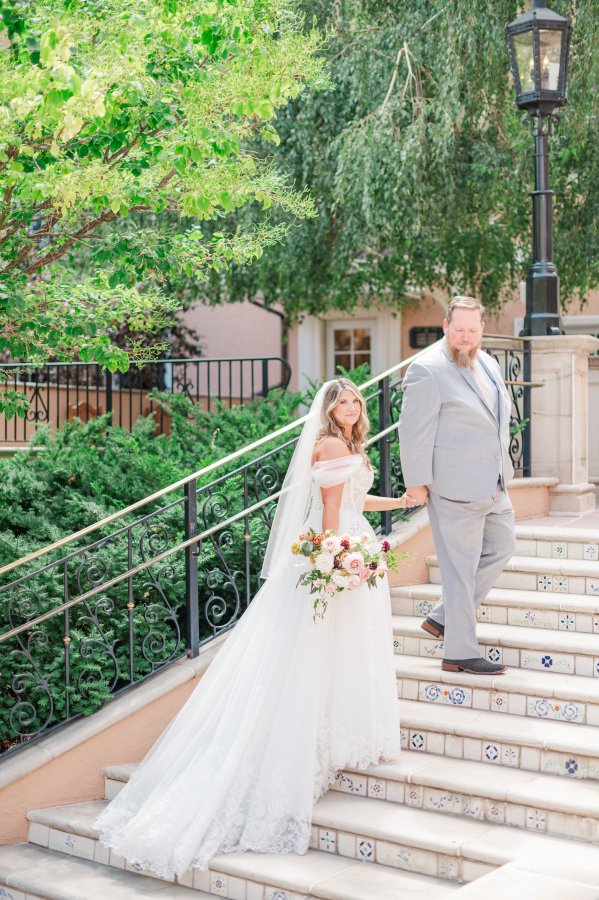 A groom leads his bride by the hand up some garden stairs as she looks back over her shoulder