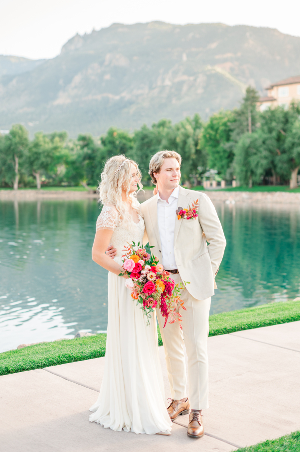 A bride and groom admire the sunset by a pond in a cream suit and lace gown with large bright bouquet