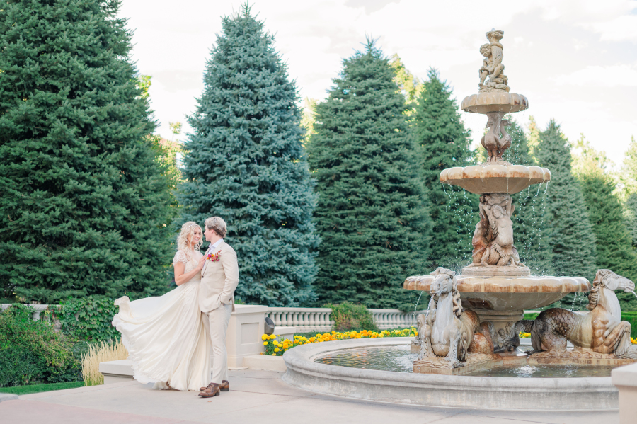 A bride and groom in tan stand by the stunning garden fountain smiling at each other at the broadmoor hotel wedding venue