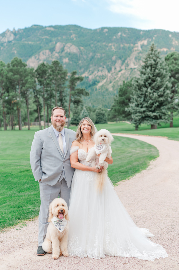 Newlyweds stand in a gravel path with their two fluffy dogs wearing custom bandanas