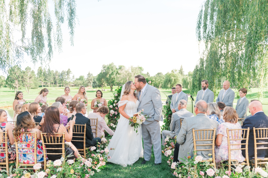 A groom in his grey suit kisses his bride in the aisle under willow trees as guests watch