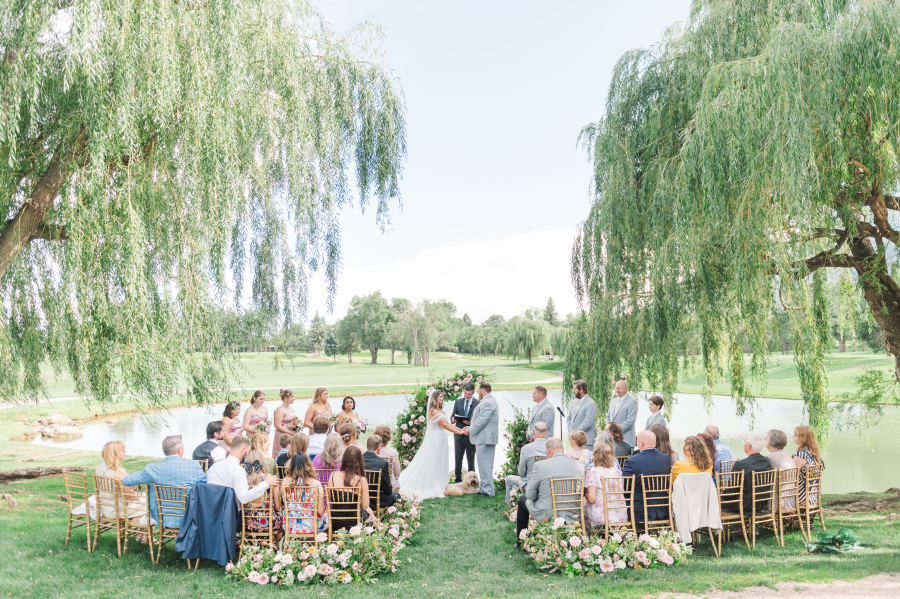 A bride and her groom in grey stand at the altar holding hands under the willow trees by the water during their broadmoor hotel wedding ceremony as guests watch
