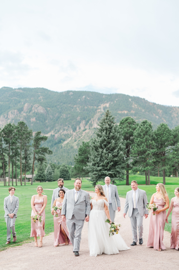 A bride and her groom in grey walk holding hands down a gravel path with their wedding party in matching pink and grey