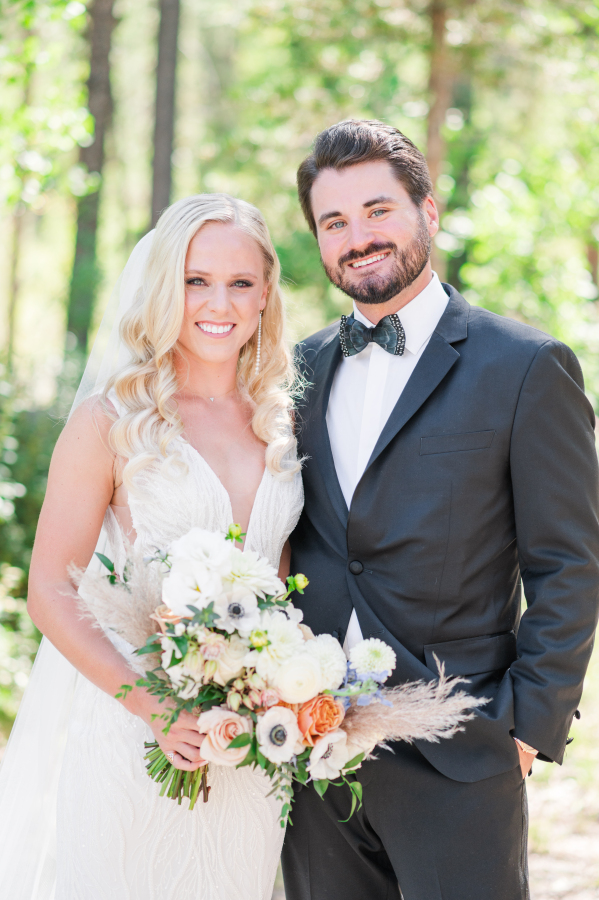 A bride and groom smile while standing in a forest holding the boho style bouquet during their camp hale weddings
