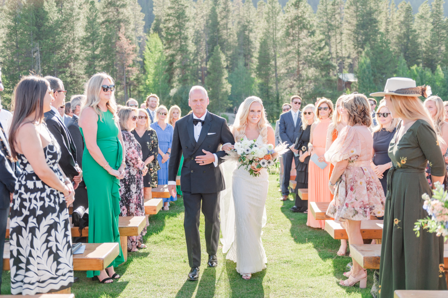 A happy bride walks down the aisle holding dad's arm to her outdoor ceremony