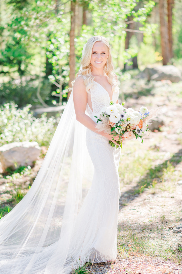 A happy bride holds her white and pink bouquet in a forest in her lace gown and long veil