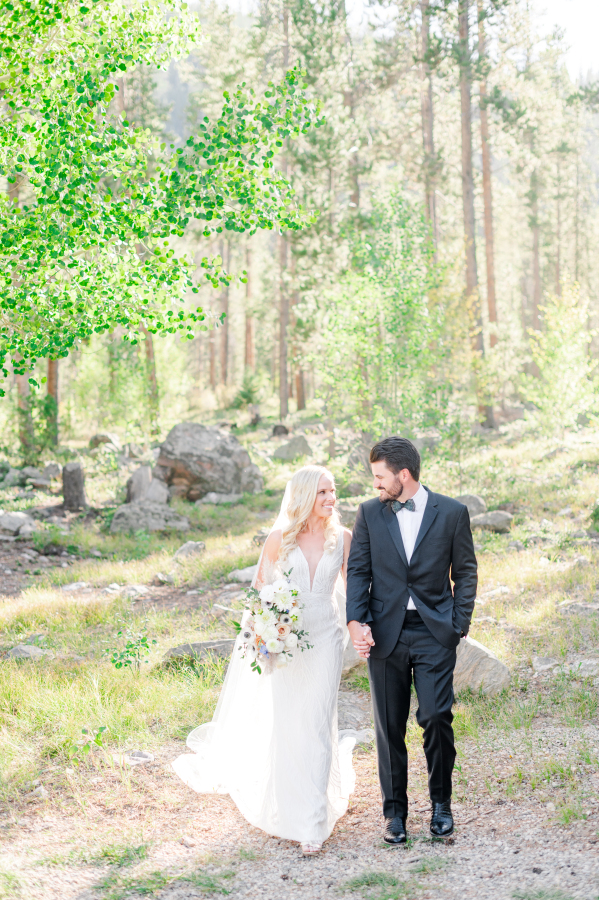 A bride and groom in his black tux walk in a forest holding hands during their camp hale weddings