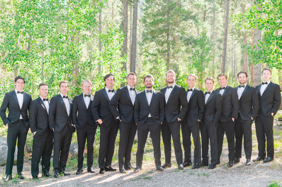 A groom stands in his black tux with his matching groomsmen on the edge of a forest with hands in pockets