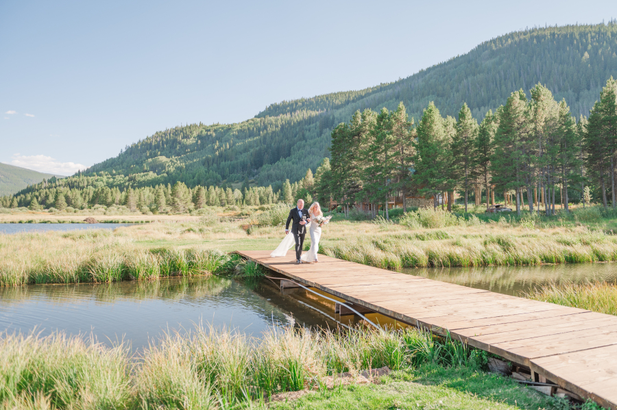 A bride walks a long dock over a pond with dad to her camp hale weddings outdoor ceremony