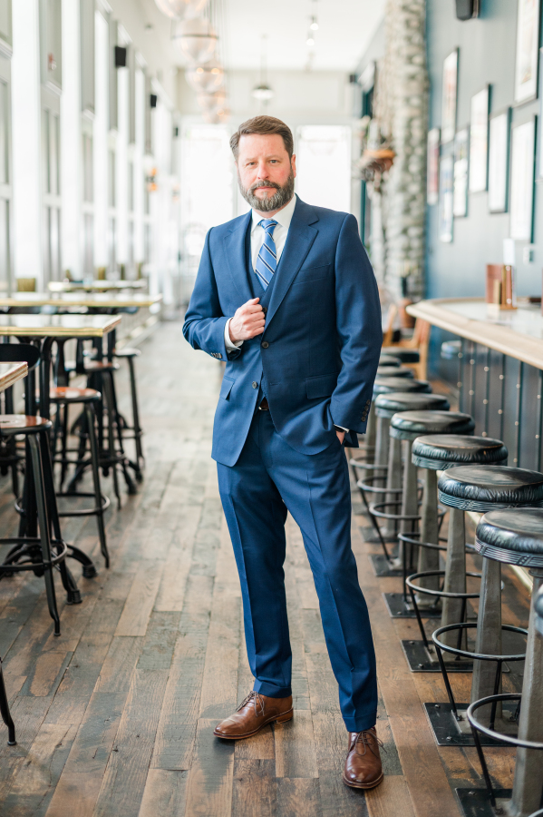 A groom in a blue suit stands with a hand on his lapel in the bar