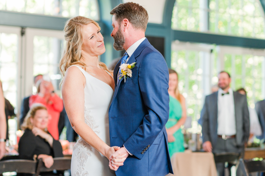A bride smiles into the eyes of her groom in a blue suit during their first dance