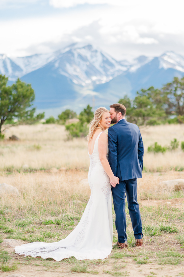 A groom whispers in his happy bride's ear while they admire the mountains and hold hands in a meadow during their surf hotel wedding