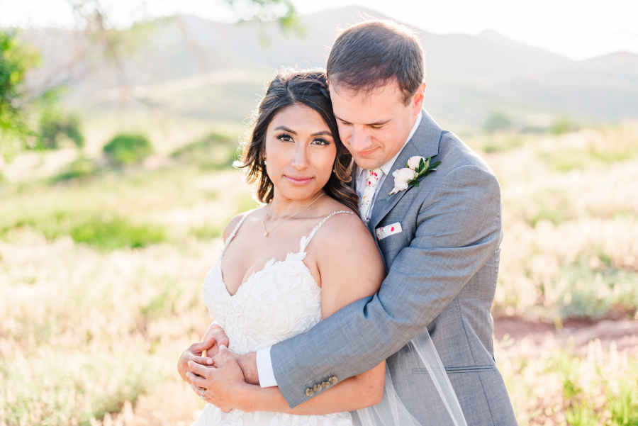 A groom in a grey suit hugs his bride from behind while standing in a meadow at sunset during their the manor house wedding