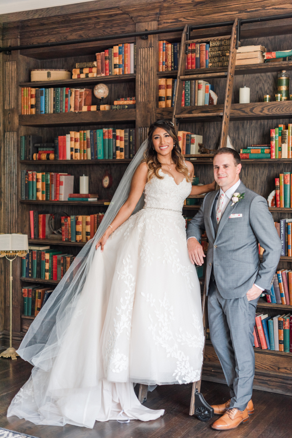A bride climbs a library ladder her groom in grey is leaning on at their the manor house wedding