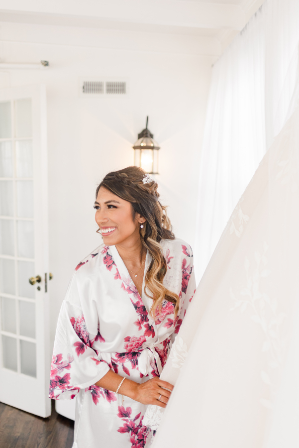 A bride in white and pink pajamas smiles big while admiring her gown hanging in a window