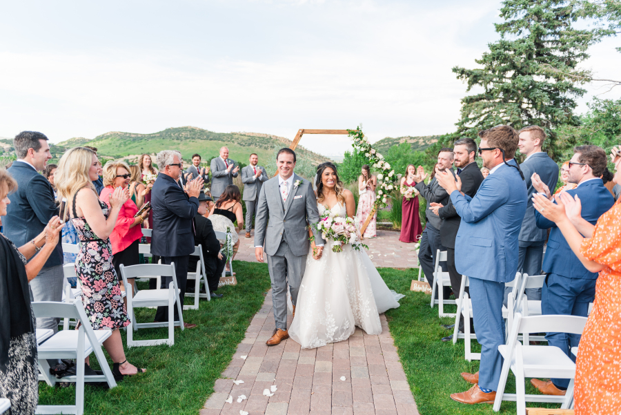 Newlyweds walk up the aisle smiling and holding hands as guests applaud during their outdoor the manor house wedding ceremony