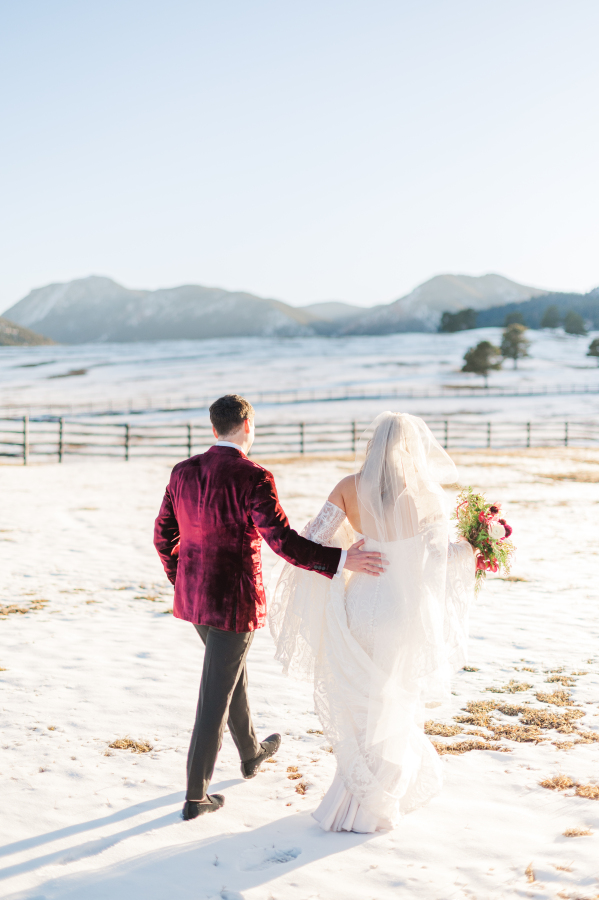 A groom in a purple velvet jacket walks his bride over snow covered pasture at sunset