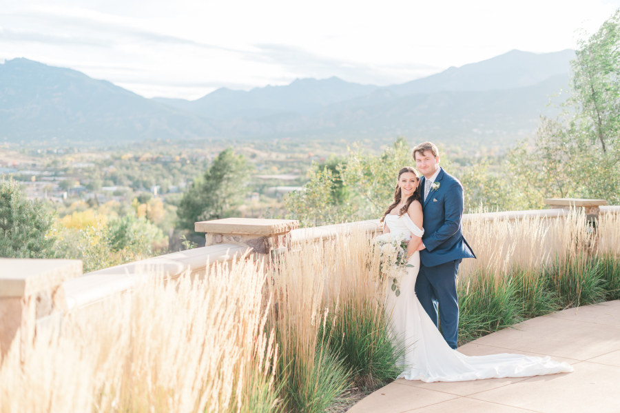 A bride and groom in a blue suit snuggle in tall grass on a patio overlook at sunset