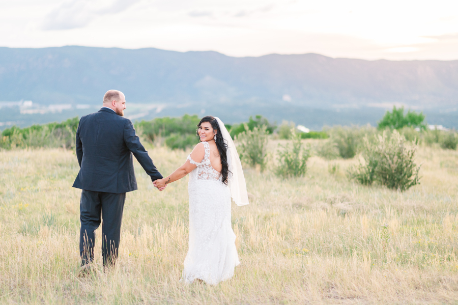 Newlyweds walk in a meadow of tall golden grass at sunset smiling over her shoulder at one of the Colorado Springs wedding venues