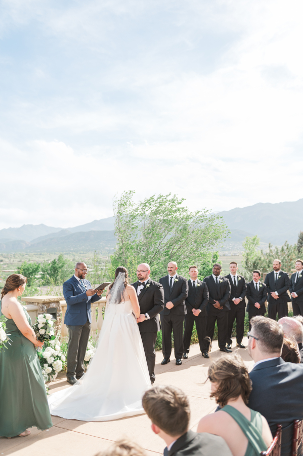 A view from an outdoor ceremony while the bride and groom hold hands listening to the officiant