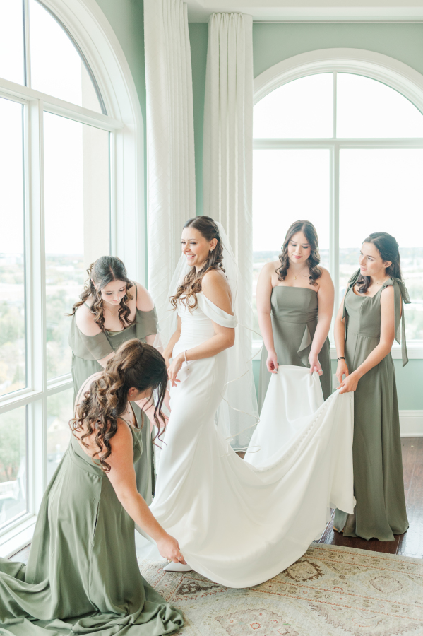 A bride stands in some windows as her bridesmaids in sage dresses help with her gown at one of the Colorado Springs wedding venues