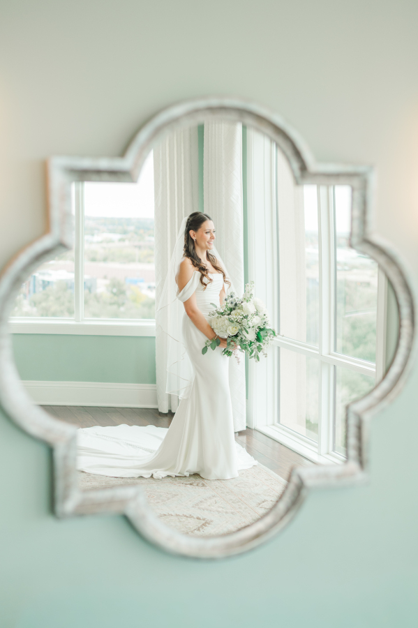 A bride stands in a window smiling as seen through a mirror