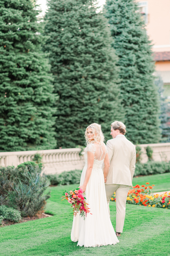 A bride and groom walk in the garden with the bride looking over her shoulder wearing lace gown with large bright bouquet
