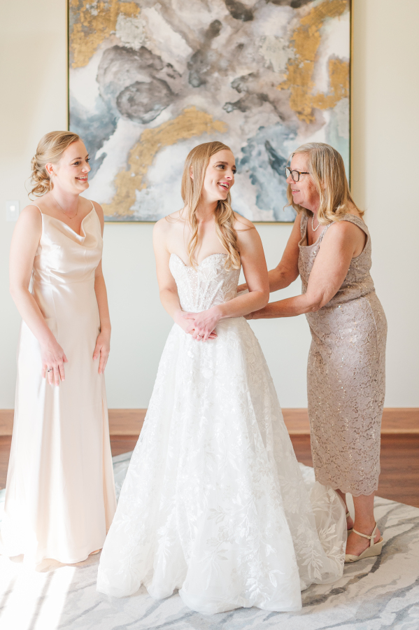 A bride laughs with her mom and sister helping get in her gown