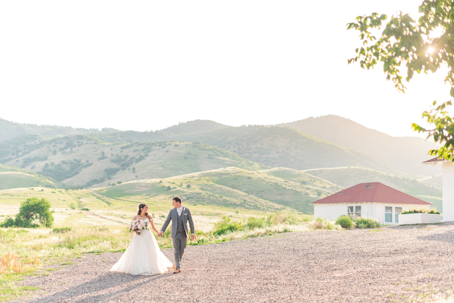 A bride and groom smile at each other while walking and holding hands at sunset in the mountains
