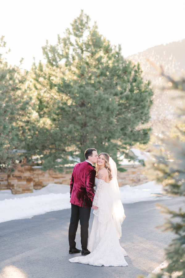A happy bride smiles while her groom in a velvet tux jacket kisses her cheek during their spruce mountain ranch wedding
