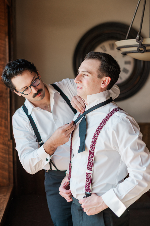 A groom gets help with his bowtie from a groomsman in the getting ready room window