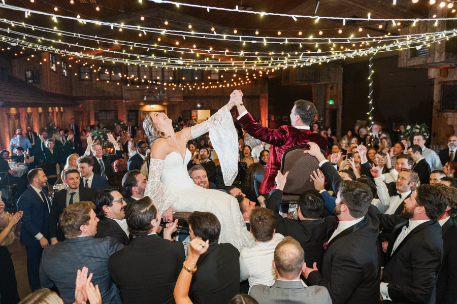 A bride and groom hold hands while being lifted in chairs by their reception guests