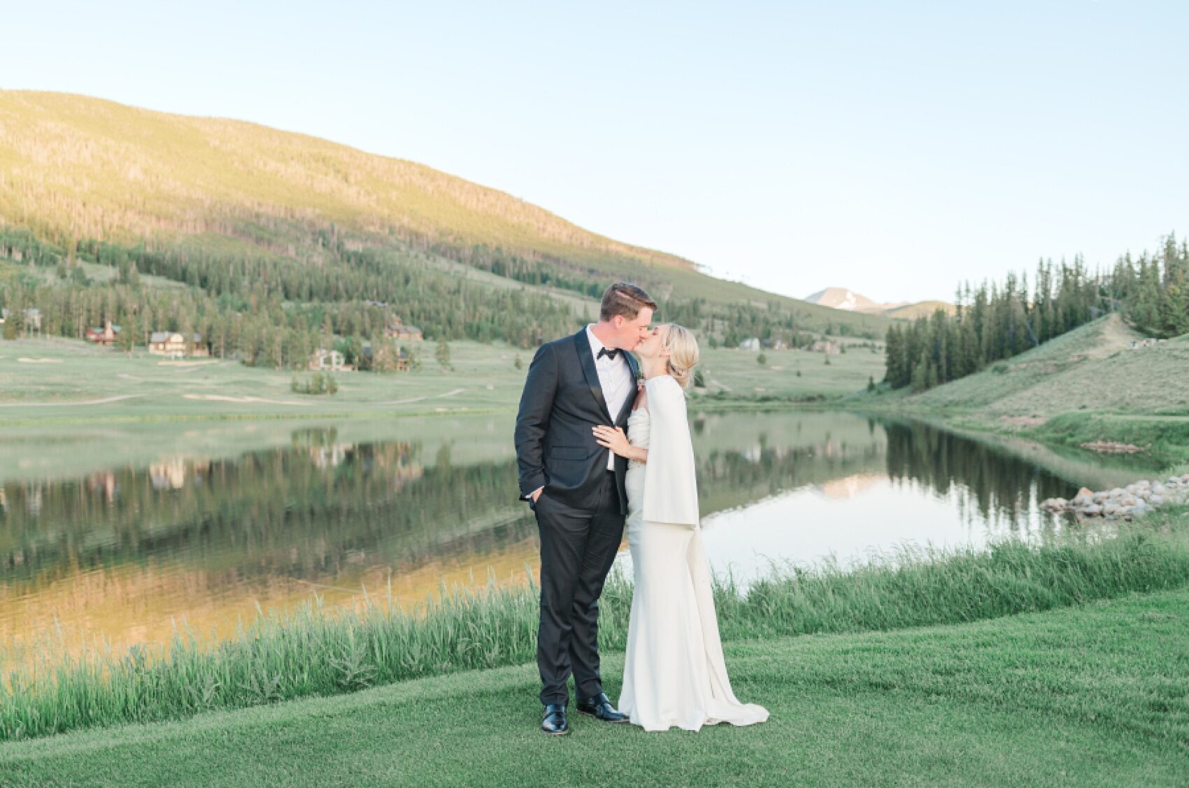 A bride and groom walk and kiss on a golf green overlooking a pond at sunset at one of the Colorado Mountain Wedding Venues