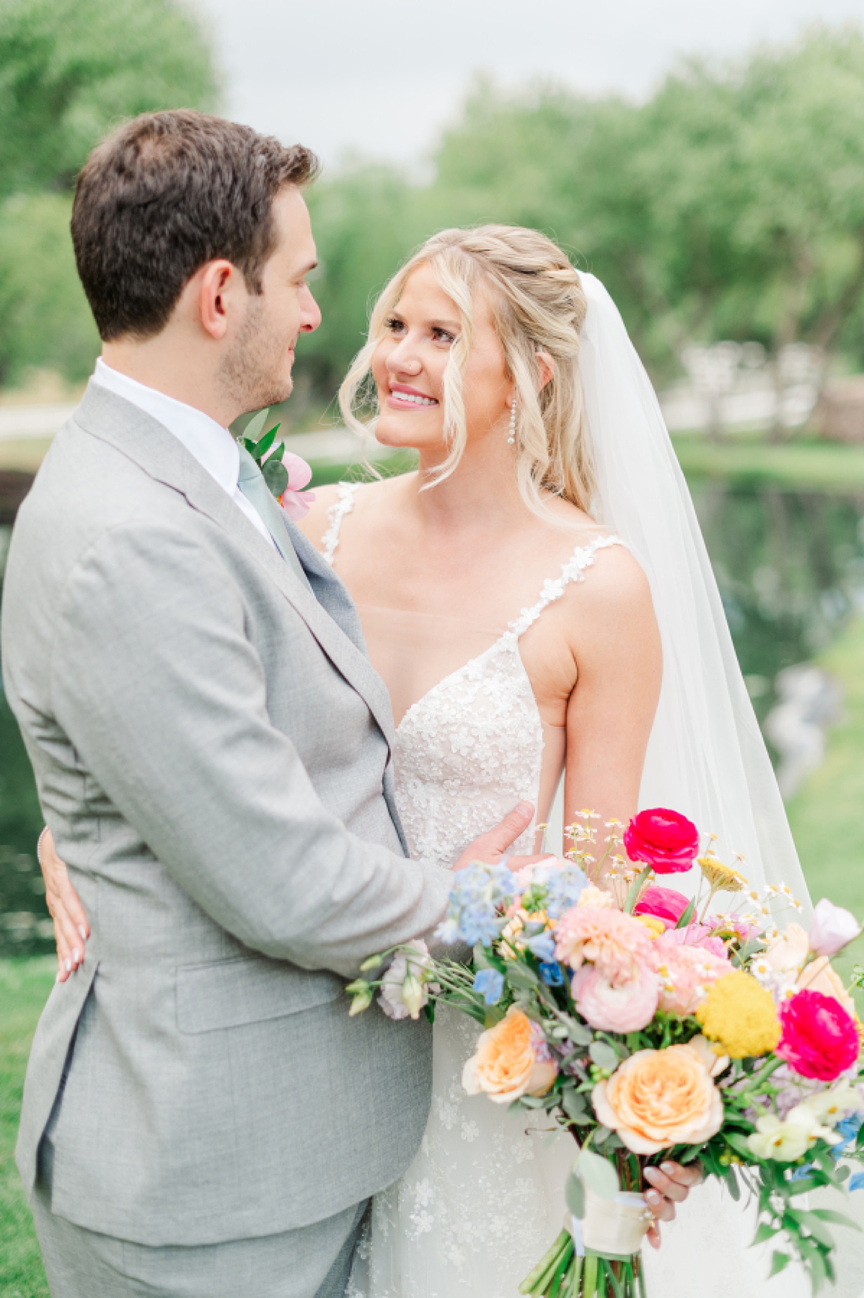 A bride and groom embrace and snuggle by a pond in lace and grey with colorful bouquet