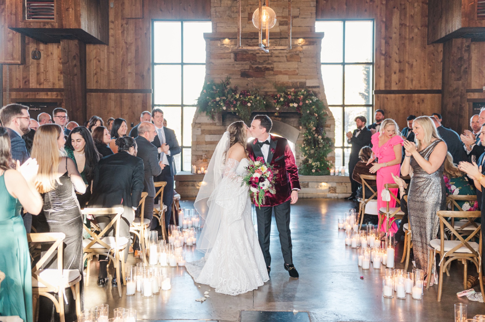A groom in a red velvet tux jacket kisses his bride in the aisle as guests clap at one of the rustic Colorado Mountain Wedding Venues