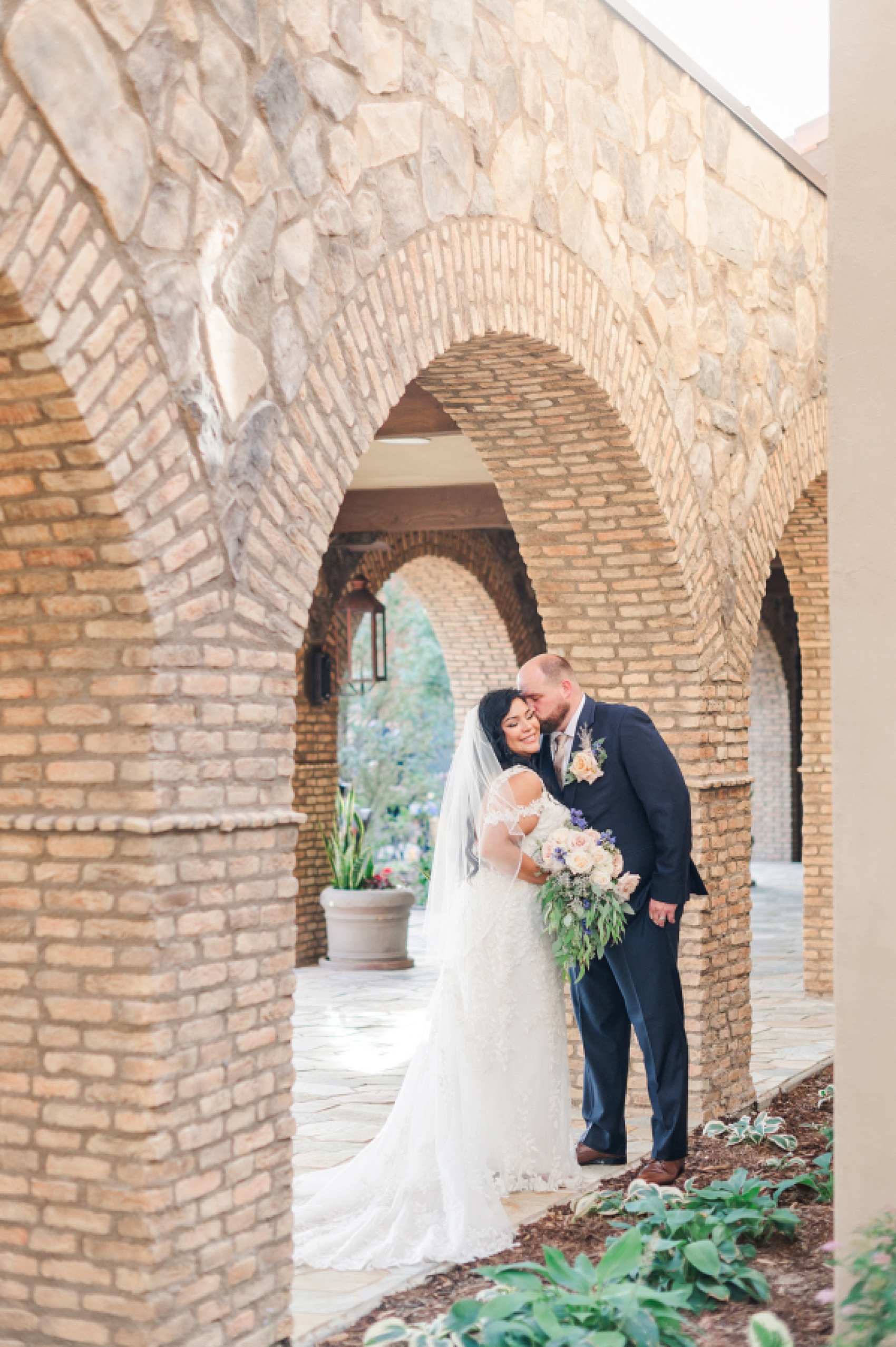 A bride smiles while being kissed on the cheek by her groom in a garden under a stone archway at one of the Denver Wedding Venues